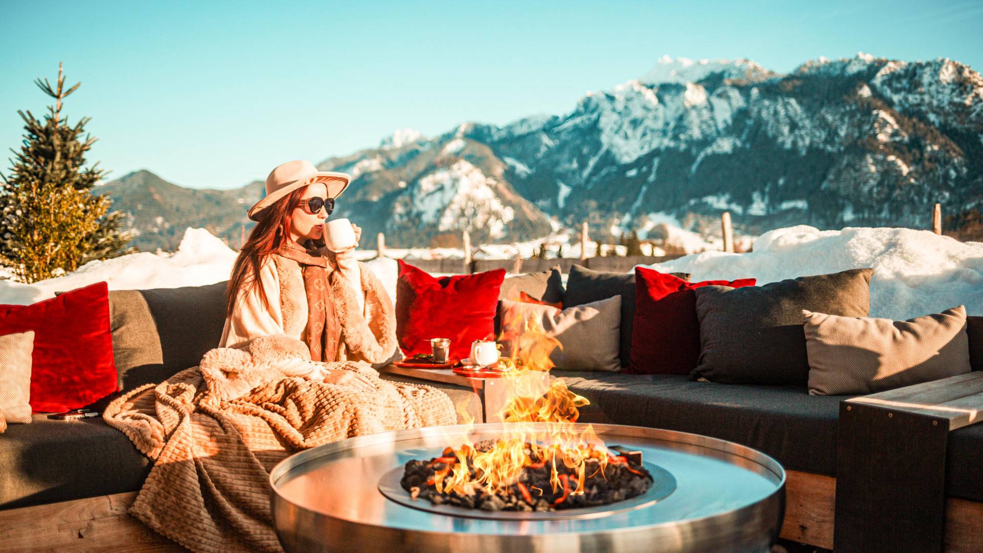 Gast entspannt mit heißem Getränk und Decke an der Feuerstelle des Rübezahl, Blick auf die verschneiten Allgäuer Berge